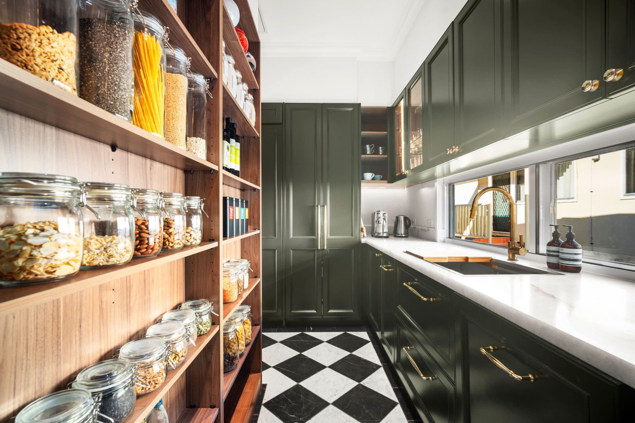 Butler’s pantry with deep green cabinetry, gold hardware, white stone benchtops, and open timber shelving.