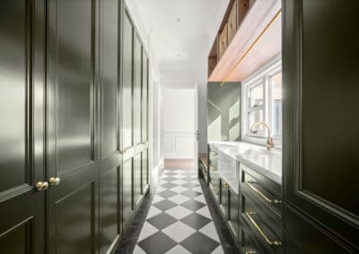 Mudroom with deep green cabinetry, gold hardware, white farmhouse sink, and checkerboard flooring.