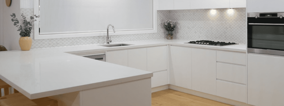 Modern white U-shaped kitchen with minimalist cabinetry, a gas cooktop, built-in oven, and under-cabinet lighting. Features a white tiled splashback with a fish scale pattern, a central sink beneath a large window, and a spacious breakfast bar with wooden stools. Decor accents include fresh flowers and a ceramic vase with eucalyptus.