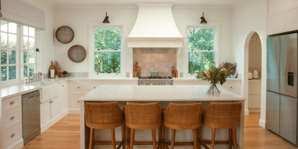 Light-filled refined country kitchen with a central island, woven bar stools, white cabinetry, timber flooring, and greenery visible through double windows.