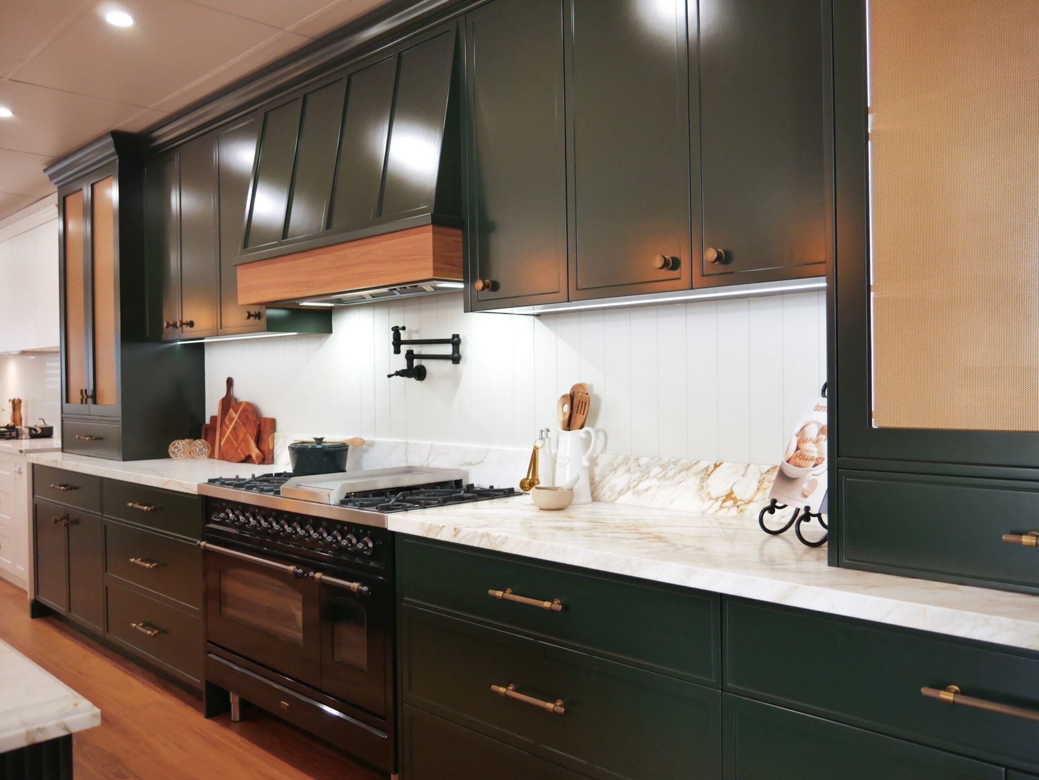 Dark green kitchen with marble island, brass fixtures, pendant lights, and velvet bar stools on timber flooring.