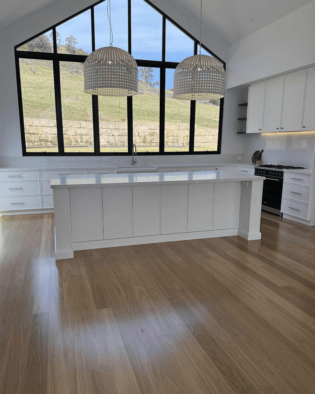 Modern country kitchen in Picton with vaulted ceilings, black-framed windows, white island benchtop, pendant lights, timber floors, and custom white cabinetry.