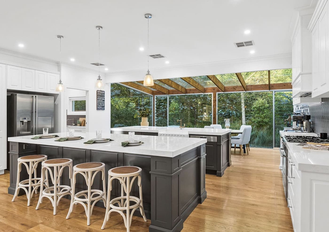 Modern open-plan kitchen design featuring a large island with dark cabinetry, white stone benchtops, natural timber floors, and floor-to-ceiling glass windows overlooking a lush garden.