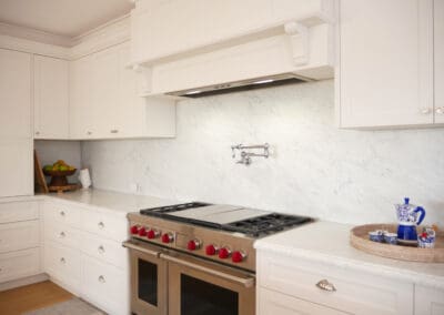 Hamptons-style kitchen featuring a stainless-steel double oven with red knobs, white shaker cabinetry, and a marble splashback with a pot filler tap.