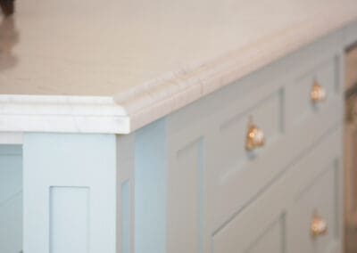 Close-up of a Hamptons-style kitchen island featuring a marble benchtop with a detailed edge profile and soft blue shaker cabinetry with brass hardware.