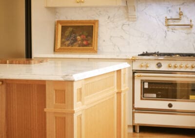 Corner detail of handcrafted timber kitchen island with decorative joinery and marble benchtop, gold freestanding range oven and marble splashback in background