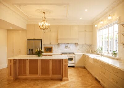 Full view of heritage-style kitchen in Croydon Park Sydney featuring ornate plaster ceiling, crystal chandelier, handcrafted timber island, cream Waratah-profile cabinetry, marble splashback and herringbone timber floors
