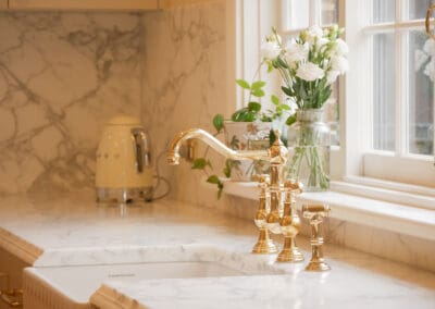 Gold bridge mixer tap and white undermount sink positioned beneath a multi-pane heritage window with potted plants and white flowers in Sydney kitchen renovation