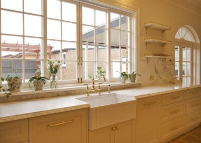 Wide view of heritage kitchen sink wall featuring apron front butler sink, polished gold bridge mixer, marble benchtop, open brass shelving and large multi-pane windows in Croydon Park Sydney