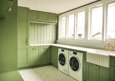 Wide view of heritage laundry in Croydon Park featuring green custom cabinetry, appliances, butler sink with gold bridge mixer, marble benchtop, beadboard panelling and large windows