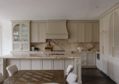 Full view of a bespoke classic kitchen featuring chiefly cabinetry, a large natural stone island, handcrafted rangehood, satin brass tapware, floor-to-ceiling cabinetry and dark herringbone timber flooring.